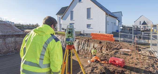 Im Vordergrund steht ein Mann in einer Warnschutzjacke, dessen Rücken zur Kamera zeigt. Er blickt auf ein Vermessungsgerät, das vor dem ausgehobenen Boden platziert ist. Im Hintergrund ist ein neu gebautes Einfamilienhaus zu sehen.