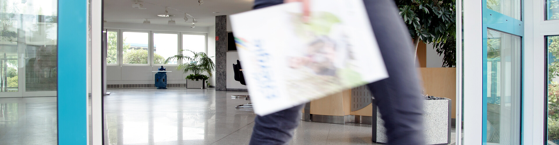 Der Foyer-Bereich hat große Fenster, einen türkisenen Türrahmen und Pflanzen. Eine Person in grauer Hose läuft leicht verschwommen durch das Bild und hält ein Kundenmagazin der Stadtwerke in der Hand.