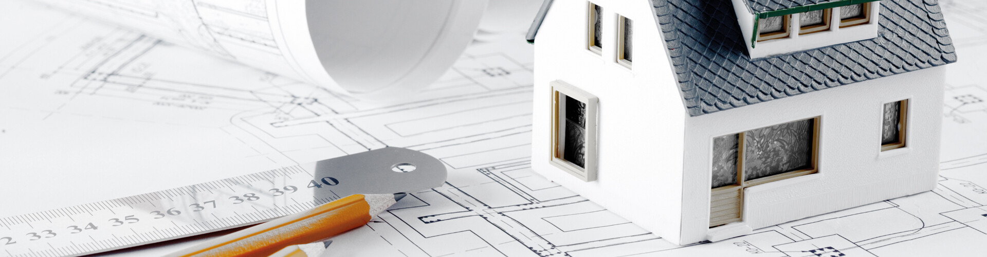Auf einem Bauplan steht ein Modellhaus. Daneben sind ein eingerollter Plan, ein Lineal und zwei Bleistifte platziert.