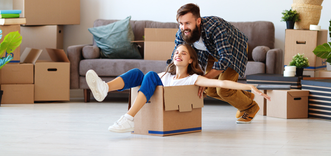 Eine lächelnde Frau sitzt mit angewinkelten Beinen in einem Umzugskarton, streckt die Arme zur Seite und grinst. Der Karton wird von einem Mann angeschoben. Im Hintergrund sind eine Pflanze, ein Sofa und mehrere weitere Umzugskartons zu sehen.