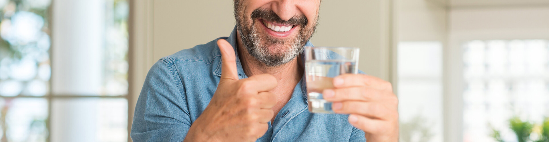 Ein Mann lächelt und zeigt den Daumen nach oben, während er in der anderen Hand ein Glas Wasser hält.