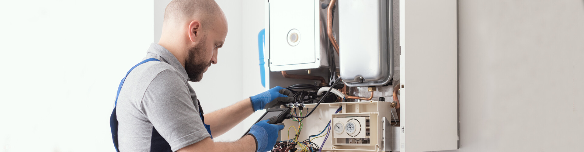 Ein Techniker in Arbeitskleidung und blauen Schutzhandschuhen führt Arbeiten an einem geöffneten Gasgerät durch. Das Gerät ist an einer weißen Wand montiert und die Abdeckung ist geöffnet, wodurch Kabel und elektronische Bauteile sichtbar sind. Der Techniker nutzt ein Messgerät, um Verbindungen und Komponenten im Inneren des Geräts zu überprüfen. 