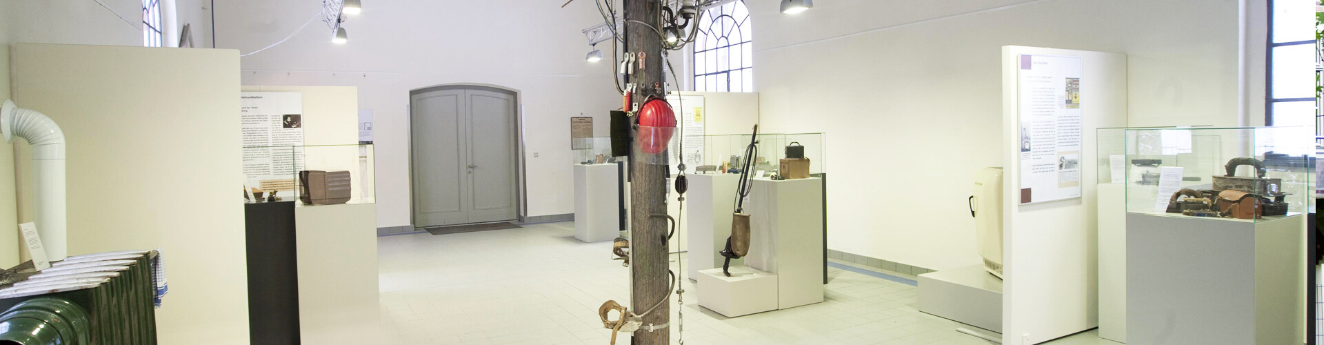 Die Innenansicht zeigt das Nebengebäude des Wasserkraftwerks „Steinerne Renne“, in dem eine kleine historische Ausstellung untergebracht ist. In der Mitte des weißen Raums befindet sich ein deckenhoher Freileitungsmast aus Holz, an dem ein Schutzhelm aufgehängt ist. An den Wänden hängen zahlreiche Informationstafeln, weitere Schilder sind im Raum verteilt. In gläsernen Vitrinen werden Ausstellungsstücke zur Geschichte der Energieversorgung präsentiert. Besonders im Fokus stehen die Anfänge der Elektrizitätsnutzung mit historischen Haushaltsgeräten und alten Energieanlagen.