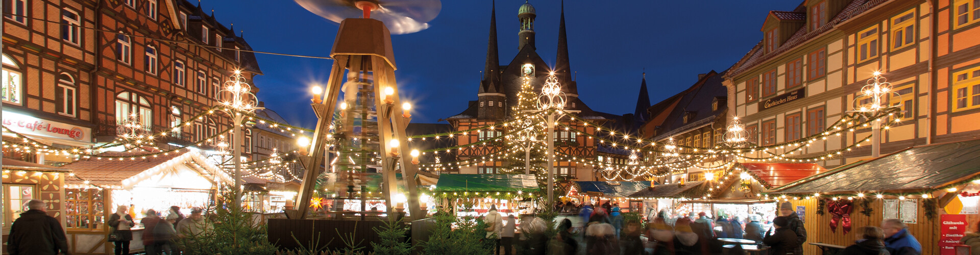 Der Wernigeröder Weihnachtsmarkt zeigt sich in der Dämmerung mit zahlreichen Buden und funkelnden Lichtern. Im Mittelpunkt steht das historische Rathaus, umgeben von Fachwerkhäusern. Erleuchtete Lichterketten schmücken den Marktplatz. 