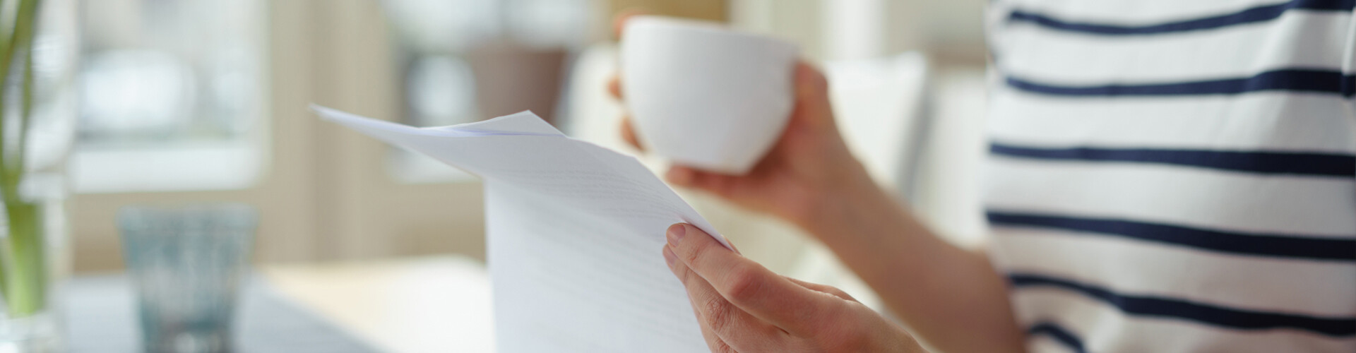 Zu sehen ist der Oberkörper einer Person in einem gestreiften Oberteil. Sie hält eine Tasse in der einen und zwei Blätter Papier in der anderen Hand.