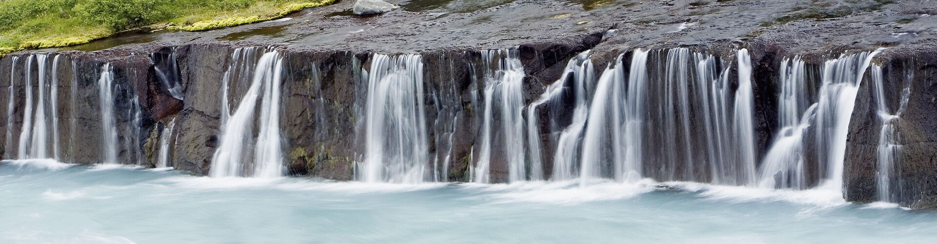 Ein langer, moosbedeckter Felsen, von dem Wasser in einem breiten Wasserfall herabfließt. Unten im Bild sammelt sich das Wasser.