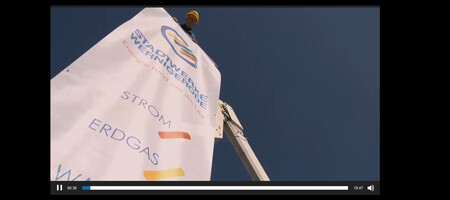 Die Aufnahme von unten zeigt eine weiße Fahne mit dem Logo der Stadtwerke Wernigerode GmbH. Ein Mann auf einer erhöhten Plattform trägt einen gelben Helm und hält die Fahne fest. Auf dem Banner sind außerdem die verschiedenen Produkte der Stadtwerke mit den jeweiligen Farben abgebildet.