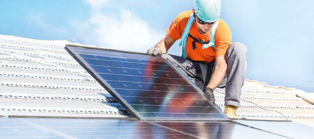 Ein Mann mit Helm und orangefarbenen Oberteil hockt auf einem weißen Dach. Er trägt Handschuhe und hält ein Photovoltaikmodule in den Händen, dass er gerade montieren möchte. Einige Module wurden bereits installiert.