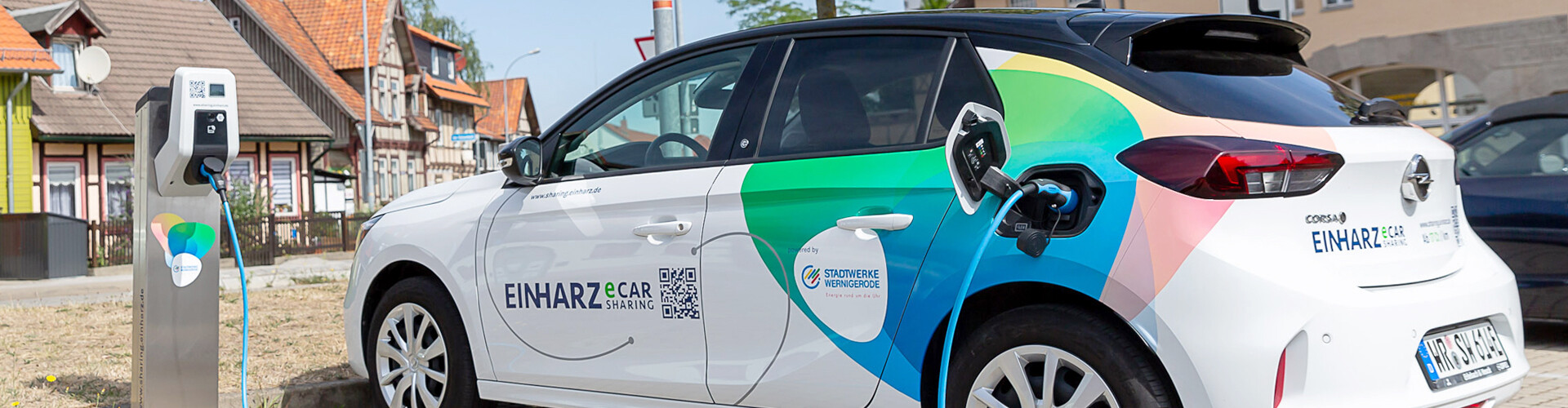 Ein e-carSharing-Fahrzeug der Stadtwerke Wernigerode GmbH lädt an einer Ladesäule. Der Wagen ist weiß, mit schwarzem Dach und Außenspiegeln, und trägt am Heck blau-grüne Designelemente. Auf der Seite befinden sich die Aufschrift „EINHARZ e-carSharing“, das Logo der Stadtwerke sowie ein QR-Code. Im Hintergrund sind Fachwerkhäuser zu erkennen.