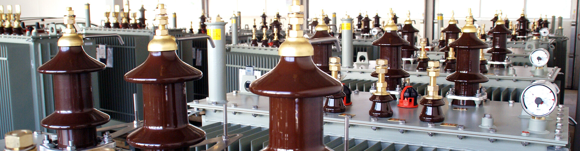 Mehrere Mittelspannungs-Transformatoren mit braunen Keramik-Isolatoren und goldfarbenen Verschraubungen in einer Industriehalle. Die Geräte sind eng nebeneinander aufgestellt.