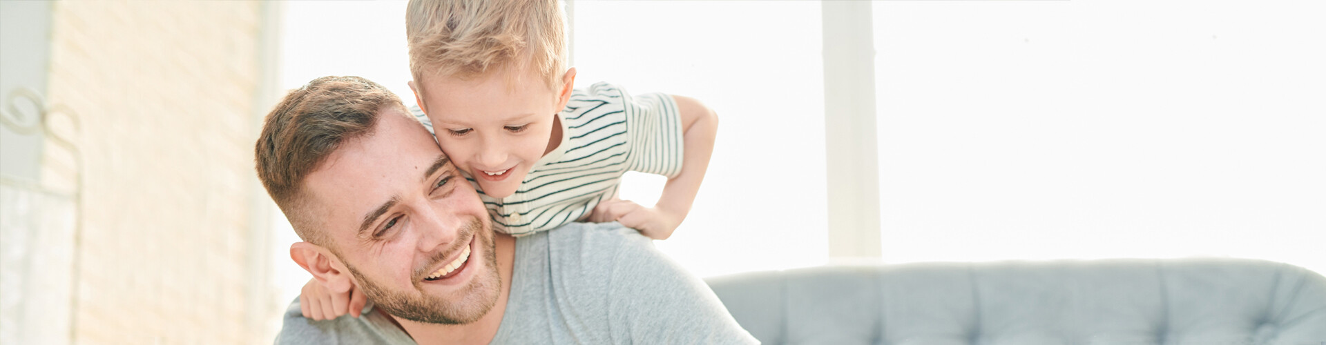 Vor hellen Hintergrund steht ein Mann mit breitem Grinsen, der den Kopf leicht zur Seite geneigt hat. Auf seinem Rücken sitzt ein kleiner Junge, der sich an seiner Schulter abstützt. Das Kind blickt nach unten und lächelt ebenfalls.