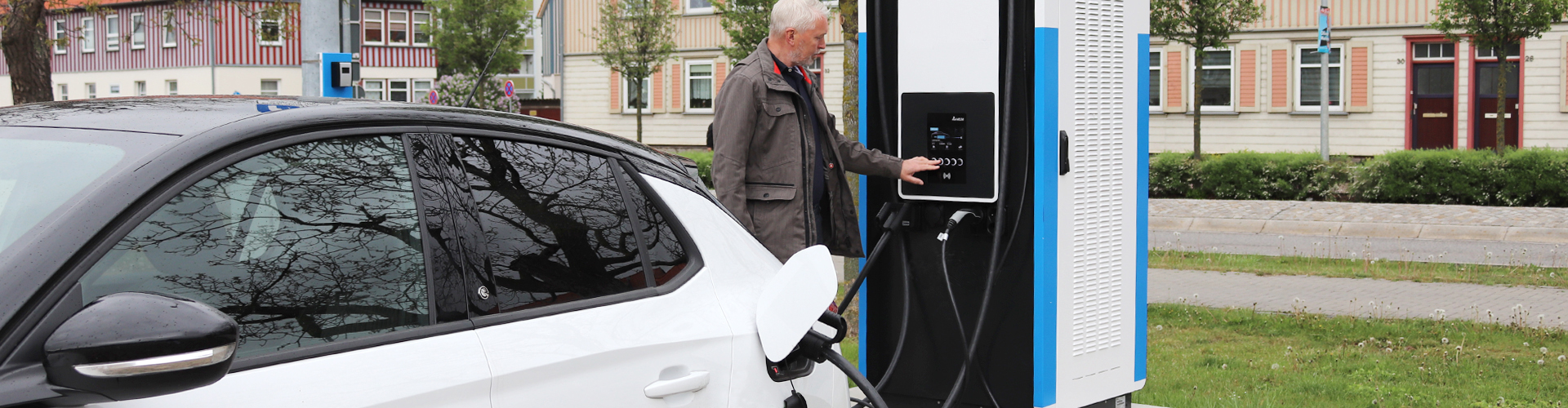 Ein weißes Elektroauto mit schwarzem Dach wird an einer Ladestation aufgeladen. Die Person bedient gerade die schwarz-weiße Ladesäule. Hinter der Station erstreckt sich eine Straße mit Häusern.