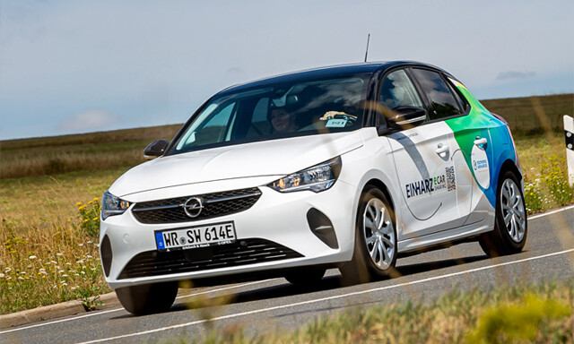 Das e-carSharing Auto der Stadtwerke Wernigerode GmbH fährt auf einer ländlichen Straße. Es ist weiß mit schwarzem Dach und hat am Heck eine blau-grüne Verzierungen. Auf der Seite hat es die Aufschrift „EINHARZ e-carSharing“, das Logo der Stadtwerke Wernigerode GmbH und einen QR-Code.