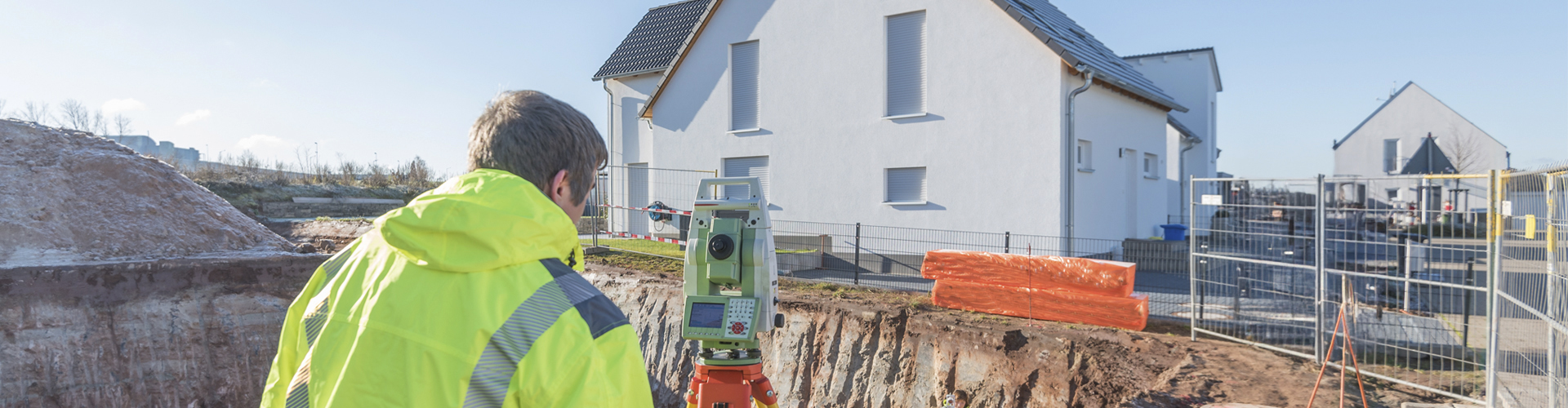Im Vordergrund steht ein Mann in einer Warnschutzjacke, dessen Rücken zur Kamera zeigt. Er blickt auf ein Vermessungsgerät, das vor dem ausgehobenen Boden platziert ist. Im Hintergrund ist ein neu gebautes Einfamilienhaus zu sehen.