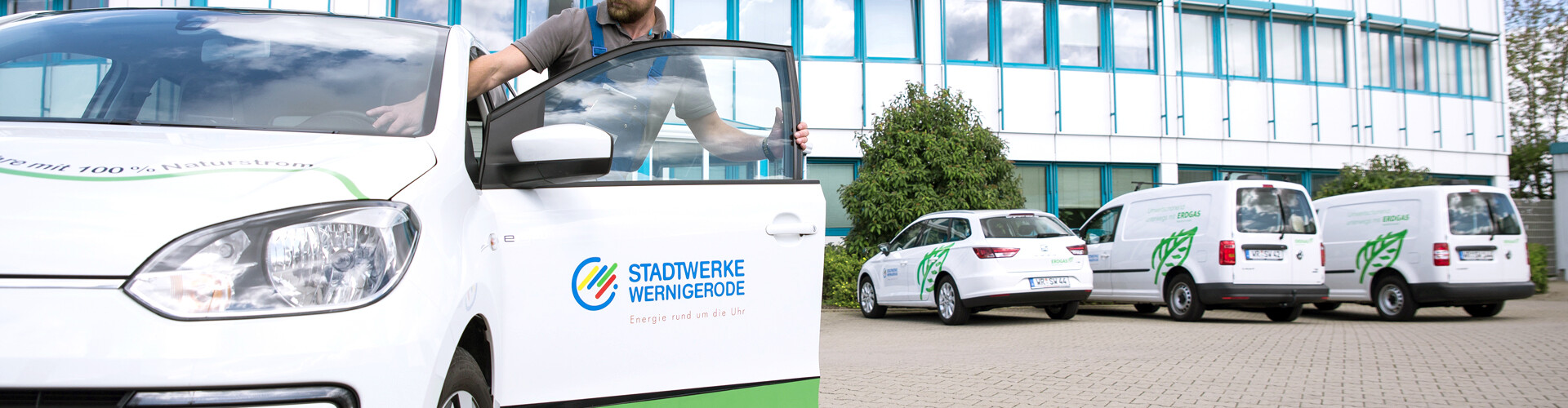 Drei weiße Firmenfahrzeuge der Stadtwerke Wernigerode GmbH stehen geordnet nebeneinander vor dem Verwaltungsgebäude. Die Fahrzeuge sind mit dem Logo, einem grünen Blatt und einem Schriftzug versehen. Auf der gegenüberliegenden Seite, der Kamera zugewandt, steht ein weiteres Fahrzeug, in das gerade ein Mann mit Arbeitskleidung einsteigt.