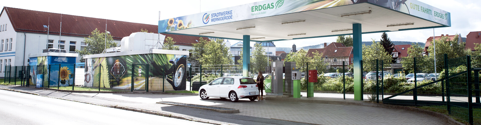 Eine Frau tankt ihr weißes Erdgasauto an einer Tankstelle, deren Dach mit dem Schriftzug „Erdgas“ sowie dem Logo der Stadtwerke Wernigerode GmbH versehen ist. Im Hintergrund sind hinter einem Zaun weitere parkende Autos sowie Häuser zu sehen.