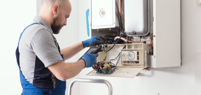 Ein Techniker führt Arbeiten an einem geöffneten Gasgerät durch. Er nutzt ein elektronisches Messgerät. Das Gerät ist an einer weißen Wand montiert und die Abdeckung ist geöffnet, wodurch Kabel und elektronische Bauteile sichtbar sind.