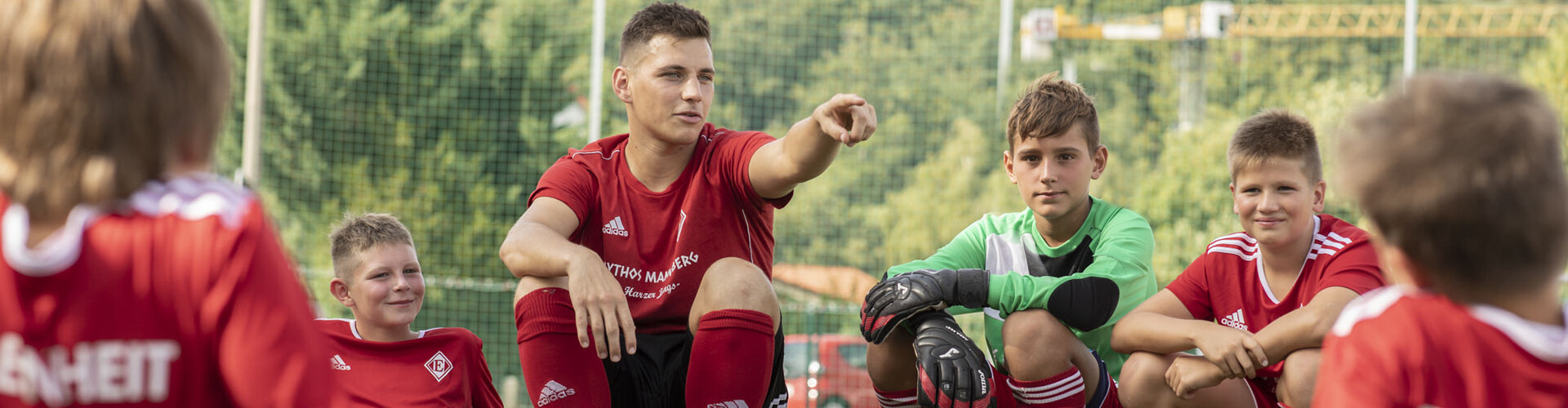 Kleine Jungen in roter Fußballkleidung sitzen mit angewinkelten Beinen auf dem Boden und stützen ihre Arme auf den Knien ab. Sie lächeln entspannt. Unter ihnen sitzt ein älterer Junge, der mit dem Finger auf etwas zeigt. Außerdem ist ein Torwart in grüner Kleidung mit Handschuhen zu sehen. Vor dem grünen Hintergrund ist ein Tornetz gespannt.