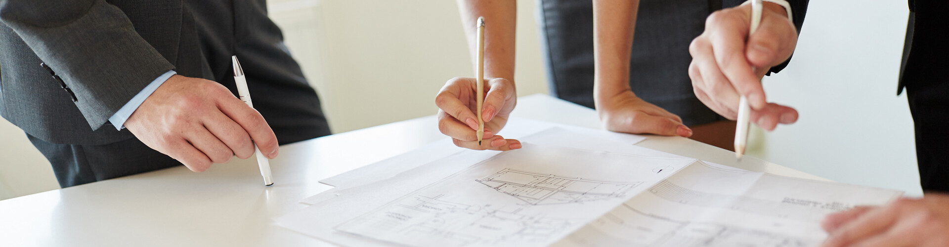   Drei Personen halten jeweils einen Stift in der Hand. Zwei von ihnen zeigen mit dem Stift auf einen Bauplan. Eine Person stützt sich mit der freien Hand an der Tischkante ab.
