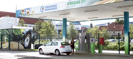 Eine Frau tankt ihr weißes Erdgasauto an einer Tankstelle, deren Dach mit dem Schriftzug „Erdgas“ sowie dem Logo der Stadtwerke Wernigerode GmbH versehen ist. Im Hintergrund sind hinter einem Zaun weitere parkende Autos sowie Häuser zu sehen.