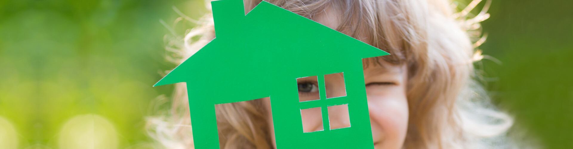 Ein Kind mit voluminösem Haar kneift ein Auge zu und blickt mit dem anderen durch das Fenster eines ausgeschnittenen grünen Papierhauses. Auch der Hintergrund ist in Grün gehalten.