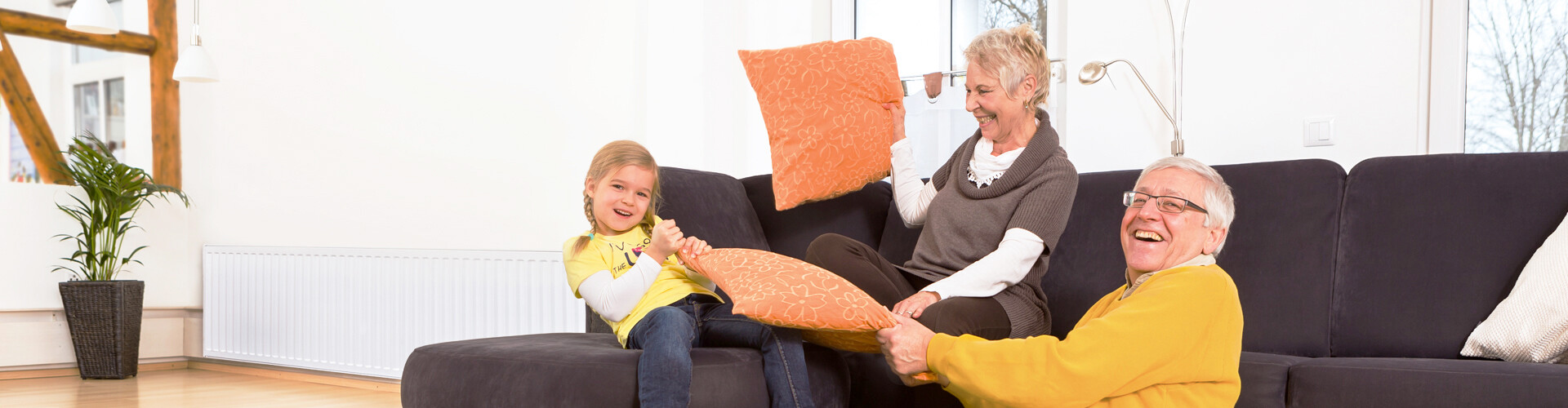 Ein Großvater im gelben Pullover und seine Enkelin im gelben T-Shirt lachen in die Kamera, während sie gemeinsam mit der Großmutter auf dem Sofa sitzen. Die Großmutter hebt spielerisch ein Kissen, als wolle sie ihre Enkeltochter damit bewerfen. Im Hintergrund sind Holzbalken und eine Pflanze zu sehen.