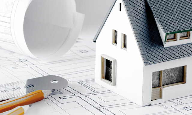 Auf einem Bauplan steht ein Modellhaus. Daneben sind ein eingerollter Plan, ein Lineal und zwei Bleistifte platziert.