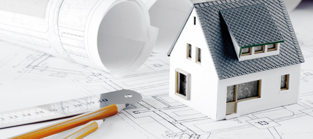 Auf einem Bauplan steht ein Modellhaus. Daneben sind ein eingerollter Plan, ein Lineal und zwei Bleistifte platziert.