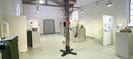 Die Innenansicht zeigt das Nebengebäude des Wasserkraftwerks „Steinerne Renne“, in dem eine kleine historische Ausstellung untergebracht ist. In der Mitte des weißen Raums befindet sich ein deckenhoher Freileitungsmast aus Holz, an dem ein Schutzhelm aufgehängt ist. An den Wänden hängen zahlreiche Informationstafeln, weitere Schilder sind im Raum verteilt. In gläsernen Vitrinen werden Ausstellungsstücke zur Geschichte der Energieversorgung präsentiert. Besonders im Fokus stehen die Anfänge der Elektrizitätsnutzung mit historischen Haushaltsgeräten und alten Energieanlagen.