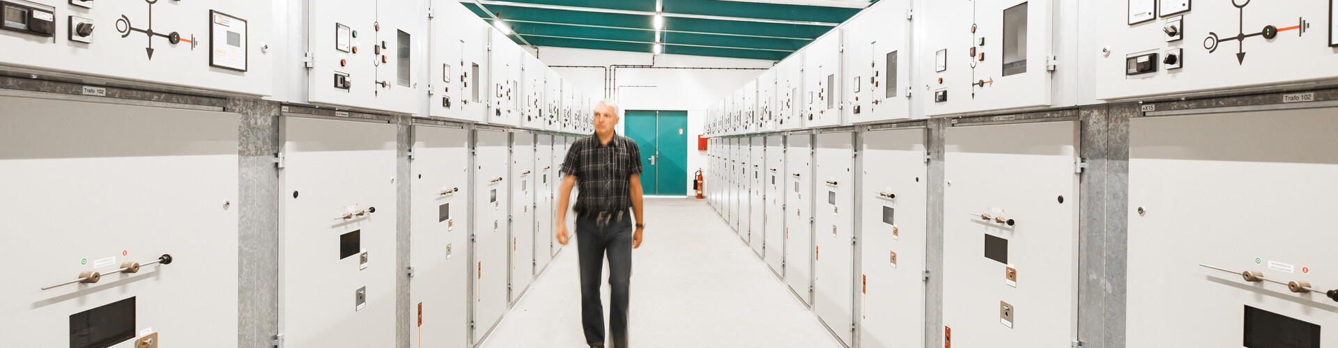 Ein Mann, bekleidet mit einem kurzärmeligen, karierten Hemd und Jeans, geht durch einen Gang innerhalb eines elektrischen Schaltanlagenraums. Links und rechts befinden sich mehrere Reihen von weißen Schaltschänken mit Kontrollleuchten, Anzeigen, Verriegelungen und Sicherheitssymbolen. Im Hintergrund ist eine türkisfarbene Doppeltür sichtbar.